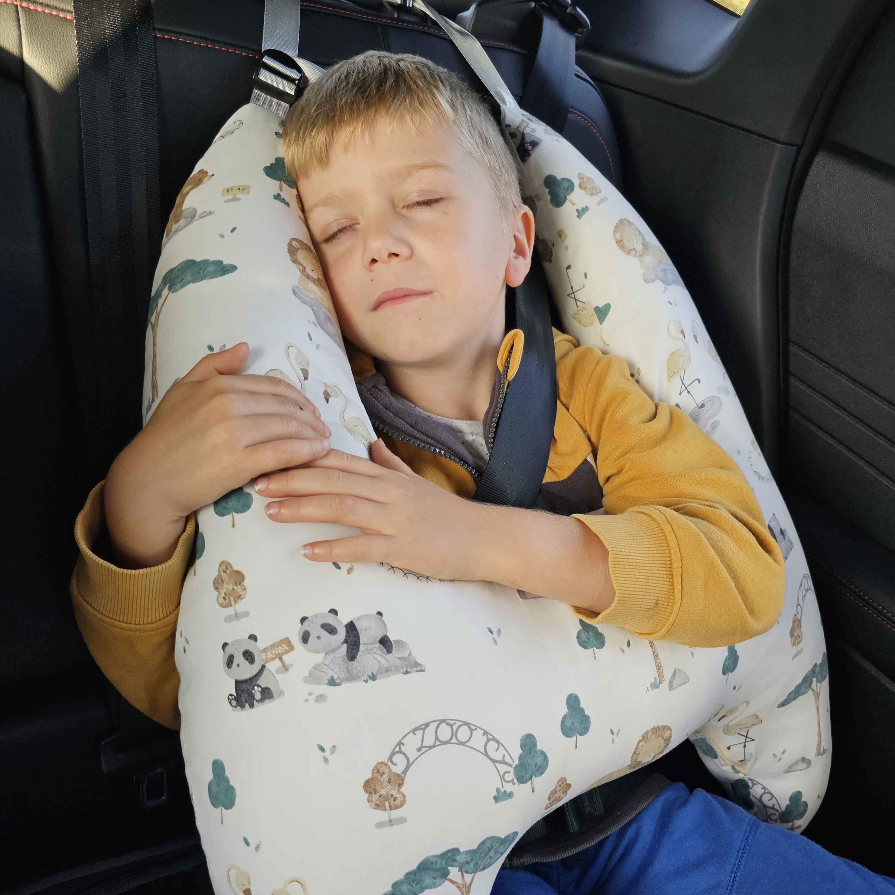 Ein schlafendes Kind im Auto, gesichert durch einen Sicherheitsgurt und gestützt durch ein großes Nackenkissen mit niedlichem Tiermuster. Ideal für Beiträge zu sicherem Kinderschlaf im Auto.