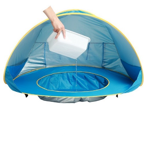Eine Hand gießt Wasser aus einem weißen Eimer in den eingebauten Mini-Pool eines aufgebauten BeachPool Zeltes in Blau mit gelben Akzenten. Der Poolbereich ist durch einen höheren, wasserdichten Rand klar abgegrenzt und befindet sich im Zentrum des Zeltes.