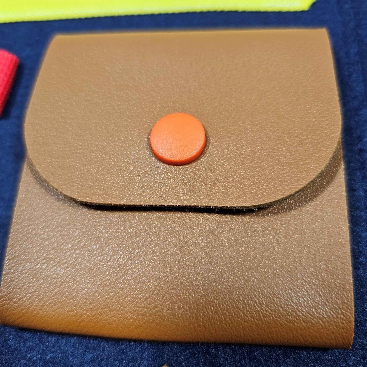 Das Bild zeigt eine Nahaufnahme eines Teils eines sensorischen Lernspielzeugs – eine braune Tasche mit einem orangefarbenen Druckknopf.